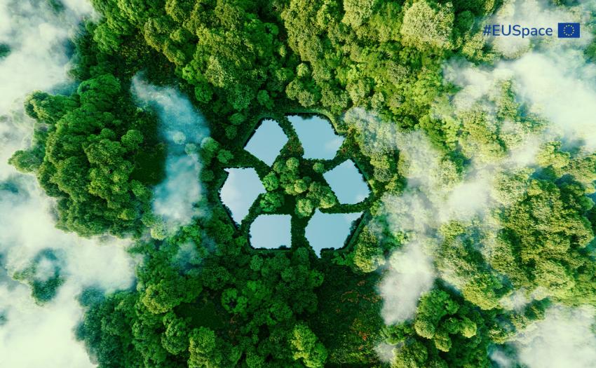 Aerial view of a dense green forest with light fog, featuring a large, stylized recycling symbol made of reflective water surfaces integrated into the landscape. In the top right corner, there is the hashtag #EUSpace alongside the European Union flag.
